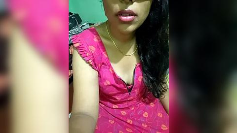 Media: Video of a South Asian woman with medium skin tone, wearing a pink dress with orange floral print, partially exposing her black bra. She has long, dark hair and wears a gold necklace. Background is blurred.