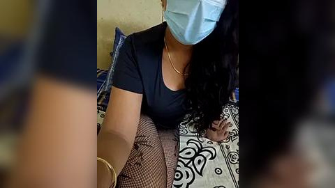 Media: Video of a woman with medium skin tone, wearing a light blue surgical mask, black top, and patterned skirt, sitting on a bed with black and white bedding.