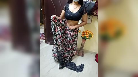 Media: Video of a woman wearing a black top and floral saree, kneeling on a tiled floor, holding the saree. Background shows a maroon door, yellow wall, and a bouquet of flowers.