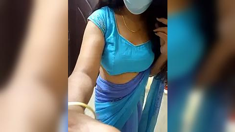 Media: A video shows a woman with medium skin tone, wearing a blue saree, mask, and a yellow bracelet, taking a selfie in a blurred background.