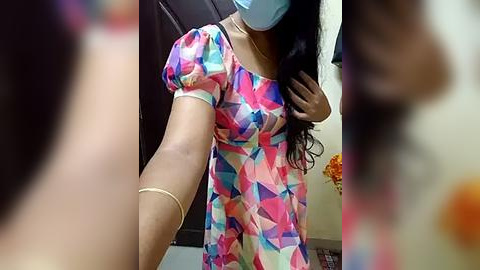 Media: Video of a woman with medium brown skin and long black hair, wearing a colorful geometric-patterned dress and a face mask, taking a selfie indoors.