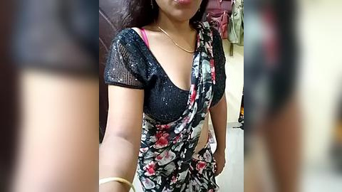 Media: Video of an Indian woman with medium brown skin, wearing a black floral sari with red and green accents, and a pink bra visible under the sari. She has a gold bracelet on her right wrist. Background shows a blurred, indoor setting.