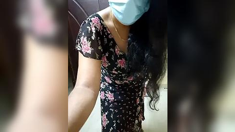 Media: A video of a woman with medium skin tone, wearing a blue surgical mask, black floral dress, and long wavy black hair. She is indoors, likely in a hospital room, with blurred background.