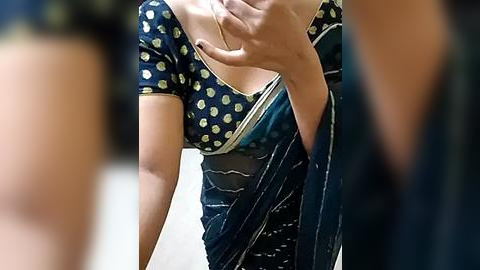 Media: A video of a woman wearing a navy blue sari with a yellow polka-dot blouse, holding her chest, partially revealing a white bra underneath. The background is blurred, focusing on her attire.