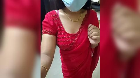 Media: Video of a woman in a red saree with gold embroidery, light skin, blue face mask, and gold necklace, taken from a slightly blurry angle.