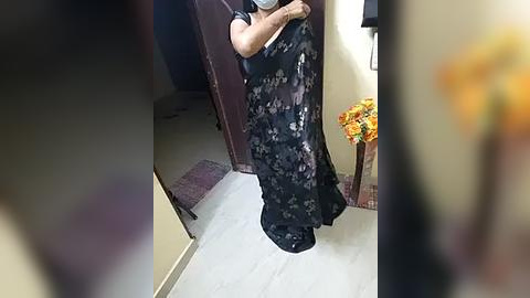 Media: A video of a person in a dark floral dress, holding a phone, standing in a room with beige walls, a carpeted floor, and a vase of flowers.