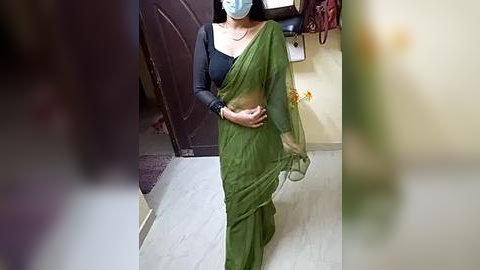 Media: Video of a woman in a green sari and black blouse, wearing a face mask, standing indoors near a closed door.