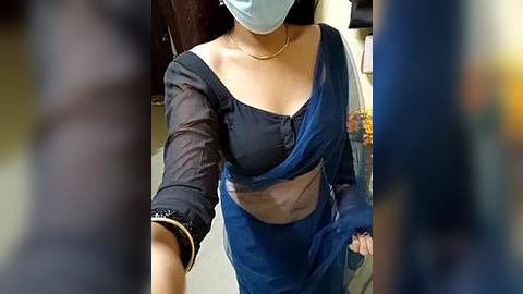 Media: Video of a woman in a blue sari, with a black blouse, blue mask, and black bracelet, walking indoors.