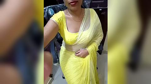 Media: Video of a South Asian woman in a bright yellow sari with a white blouse, smiling, in an indoor setting, blurred background.