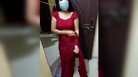 Media: Video of a South Asian woman with medium skin tone, wearing a red saree, blue face mask, and a gold bracelet. She stands in a clinic hallway with beige walls and dark wooden doors.