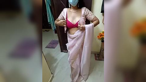 Media: Video of an Indian woman in a pink sari, blue mask, and red bra, standing in a room with a mirror, flowers, and purple mat.