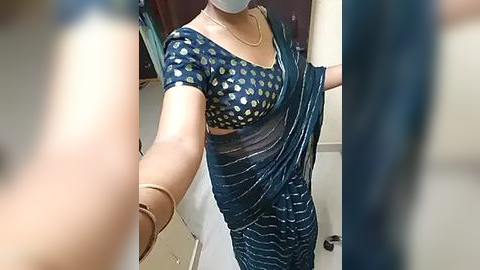 Media: Video of a woman with light brown skin wearing a blue sari with gold polka dots and a beige blouse, standing in a bathroom with a white door and a brown cabinet.