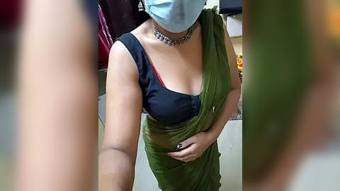 Media: Video of a South Asian woman in a green sari, black blouse, and silver necklace, wearing a blue surgical mask, taken from the waist up, indoors with blurred background.