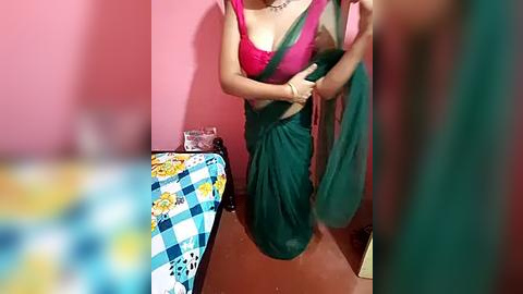 Media: A video of a woman in a green saree with a pink blouse, standing in a pink-walled room with a checkered bedspread, blurred in the foreground.