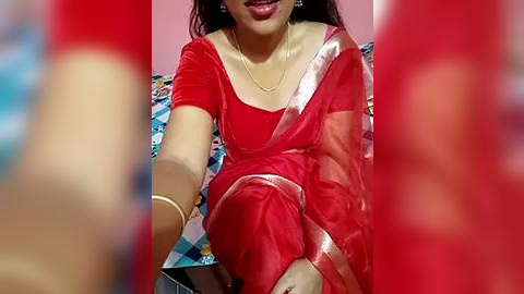 Media: Video of a South Asian woman with medium skin tone, wearing a red sari with gold trim, seated on a bed with a blue and white checkered blanket. Her face is partially visible, smiling, with dark hair.