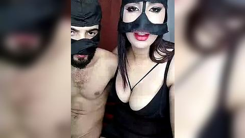 Media: Video of a couple in BDSM attire; man topless with a black mask and woman in a black mask and revealing outfit, standing indoors.