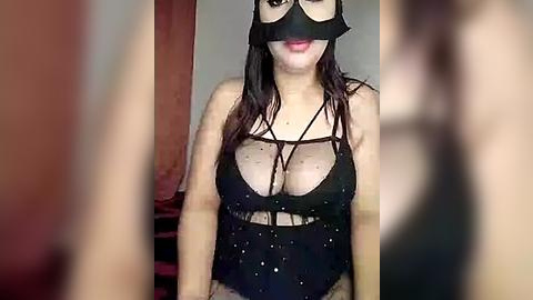 Media: Video of a woman with medium skin tone and long black hair, wearing a black lace mask, revealing a black strappy lingerie top with a cutout design. Background shows blurred red curtains and a neutral wall.