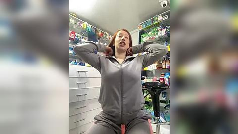 Media: A video of a woman with long red hair, wearing a grey tracksuit, stretching in a brightly-lit, cluttered room with shelves filled with colorful products.