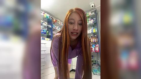 Media: Video of a young Asian woman with long, straight orange hair, wearing a purple shirt, smiling in a brightly lit convenience store. Background features shelves stocked with various products.