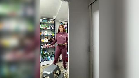 Media: Video of a woman in maroon athletic gear standing in a dimly lit room, looking at herself in a full-length mirror. Behind her, shelves are stocked with various items.