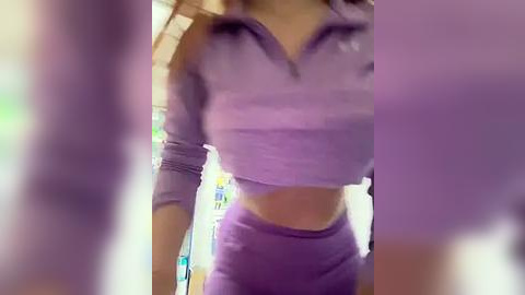 Media: A video of a woman with fair skin, wearing a purple cropped sports top and matching leggings, standing indoors with a blurred background.