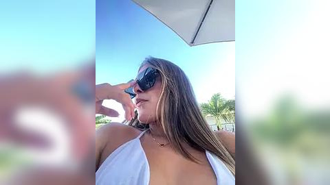 Media: Video of a tan woman with long brown hair, wearing sunglasses and a white halter top, posing outdoors under an umbrella, with blurred greenery in the background.