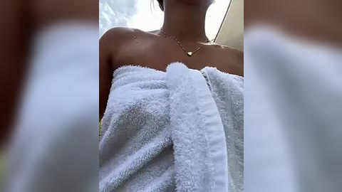 Media: Video of a young woman with light skin, wrapped in a white towel, wearing a delicate gold necklace. Background is blurred, with soft lighting.