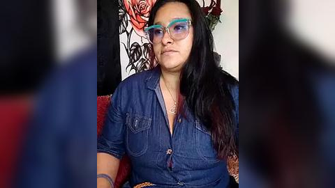 Media: Video of a middle-aged, dark-haired woman with glasses, wearing a denim shirt, sitting on a red couch. Floral-patterned wall and blurred background.