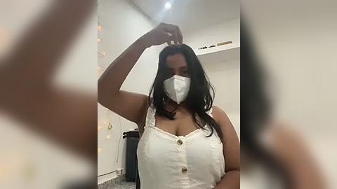 Media: Video of a woman with long black hair, wearing a white dress, white face mask, and a black face shield. She is adjusting her hair with her left hand. Background shows a white room with minimal decor.