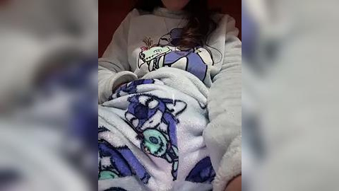 Media: Video of a person, likely a child, wearing a gray sweatshirt with a cartoon dinosaur design and a blue and white blanket featuring a blue monster. Background is blurred, focusing on the cozy, casual attire.