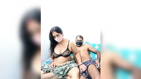 Media: Video of a young South Asian woman in a black lace bra and matching panties, sitting on a bed with a shirtless man in blue shorts, both masked, in a dimly lit room.