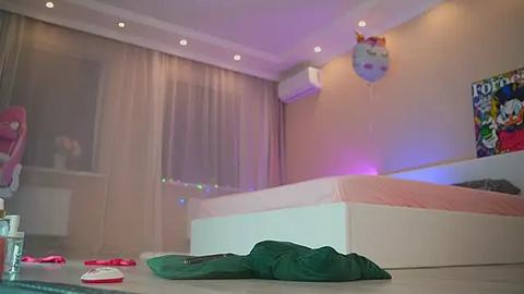 Media: Video of a modern, brightly lit bedroom with a white bed, beige walls, sheer curtains, and colorful balloons. A green bag lies on the floor near the bed.