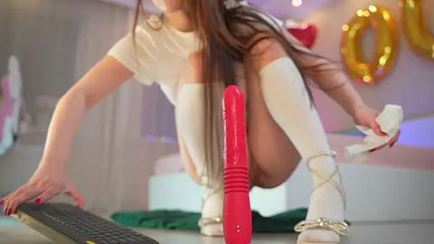 Media: Video of a young woman with long brown hair, wearing white knee-high socks, a white top, and white high heels, squatting near a red dildo and a black computer keyboard in a modern, colorful room.