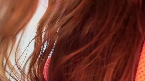 Media: Video of a close-up of a woman's shoulder-length, wavy brown hair, with strands slightly parted to reveal a red-pink streak, against a blurred background.