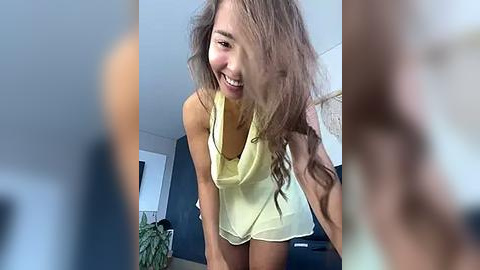 Media: A video of a smiling woman with long, wavy brown hair, wearing a yellow tank top and shorts, bending over, indoors with blurred background.