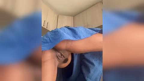 Media: A blurred video of a person with light skin and straight black hair, wearing a blue towel, lying on their side in a kitchen with beige cabinets.