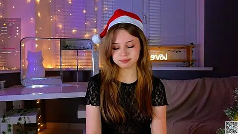 Media: Video of a young woman with long brown hair, wearing a red Santa hat and black lace top, sitting in a cozy room with string lights, a cat statue, and a \"Happy Holidays\" sign.