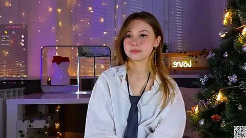 Media: Video of a young Caucasian woman with light skin and long brown hair, wearing a white lab coat over a blue top, standing in a brightly lit room with a decorated Christmas tree and a \"Lava\" sign.