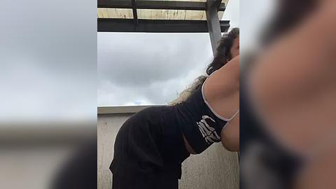 Media: Video of a woman in a black tank top and sweatpants, performing pull-ups on a metal beam under a cloudy sky.