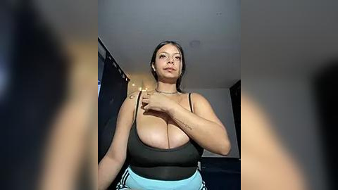 Media: A video of a Latina woman with dark hair and large breasts, wearing a black spaghetti-strap top and light blue pants, standing in a dimly lit room with black curtains and a TV in the background.