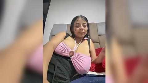 Media: Video of a Latina woman with medium skin tone, long black hair, and a medium build, wearing a pink halter top and black skirt, sitting on a bed with a gray headboard and red pillow, in a dimly lit room.