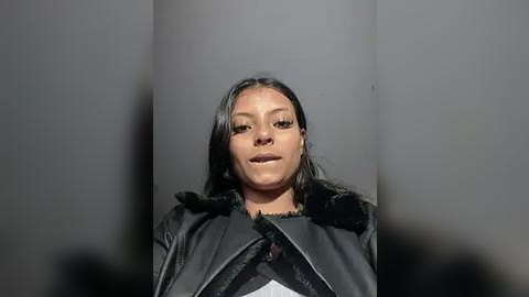 Media: Video of a woman with medium skin tone, dark hair, and a silver nose ring, wearing a black fur-trimmed leather jacket, standing in front of a gray column, looking straight at the camera.