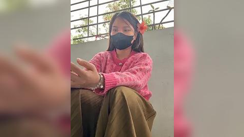 Media: Video of a young Asian woman with long black hair and a pink flower in her hair, wearing a pink sweater, khaki pants, and a black face mask, sitting on a balcony.