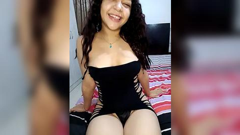 Media: Video of a smiling woman with long curly hair, wearing a black, strapless lingerie top with cutouts and black panties, sitting on a bed with a striped blanket.