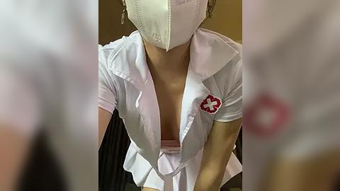 Media: Video of a female nurse in a white short-sleeved blouse, partially unbuttoned, revealing cleavage, wearing a white surgical mask. Background is blurred with a hint of a medical setting.