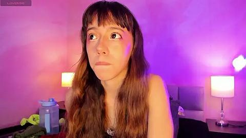 Media: A video of a young woman with long brown hair and fair skin, wearing a black top, looking contemplative against a pink and purple-lit bedroom background with two lamps and a water bottle.