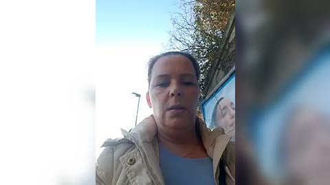 Media: Video of a middle-aged woman with light skin, brown hair, wearing a beige jacket over a blue shirt, standing outdoors in a street with a clear blue sky, blurred background.