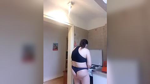 Media: Video of a woman with light skin, wearing black lingerie, standing in a bare, unfinished room with exposed walls and a white sink, viewed from behind.