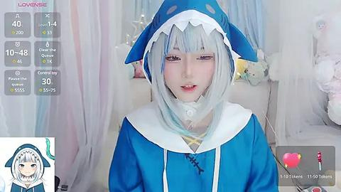 Media: A video of a young Asian woman in a blue shark cosplay costume, with a white hood and collar, against a soft, pastel-colored background. The image is overlaid with a live-streaming overlay displaying viewers' comments.