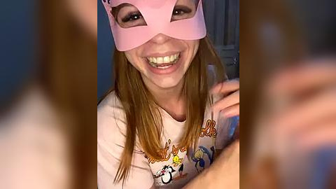 Media: Video of a smiling young woman with light skin and long straight brown hair, wearing a pink Venetian mask and a graphic t-shirt featuring cartoon characters, indoors with blurred background.